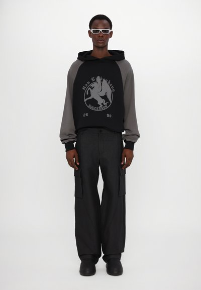 Sweat à capuche noir et gris avec un design graphique, associé à un pantalon cargo noir et des chaussures noires à enfiler. Le modèle se tient devant un fond blanc.