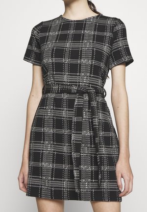 Femme portant une robe à manches courtes à carreaux noirs et blancs avec une ceinture nouée à la taille, debout devant un fond uni.