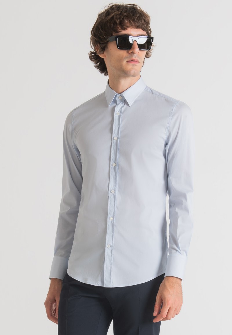 Camisa de manga larga azul claro, hecha de una tela suave, con un collar clásico y frente abotonado, combinada con gafas de sol negras.