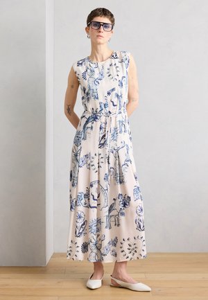 Kobieta w białej, bezrękawnikowej sukni maxi z niebieskimi abstrakcyjnymi wzorami, białe baleriny z paskiem z tyłu, krótkie włosy, okulary przeciwsłoneczne, stojąca w pomieszczeniu.