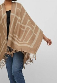 Beżowy poncho z geometrycznymi kremowymi wzorami, frędzlami i luźnym krojem. Wykonane z miękkiego materiału, narzucone na czarną bluzkę i niebieskie dżinsy.