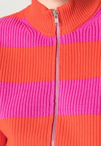 Primo piano di un maglione a zip in maglia a coste con ampie righe orizzontali rosa e arancioni e una zip argentata.