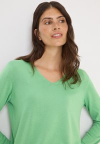 Vihreä v-pääntie neulepaita, joka on valmistettu kevyestä materiaalista ja siinä on sileä tekstuuri. Varustettu pitkillä hihoilla ja hienovaraisilla ompel yksityiskohdilla.