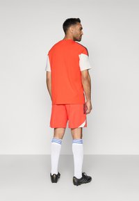 Narancssárga sportmez és rövidnadrág fehér díszítésekkel, fekete cipőkkel és kék csíkos fehér zoknikkal, hátulról nézve.