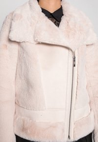 Faux fur-jacka i mjuk ljusrosa med en shearling-krage, dragkedja och texturerad yta. Har en figursydd midja och kontrasterande paneler.