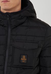 Giacca a vento nera con cappuccio, texture trapuntata, chiusura con zip frontale e taschino sul petto con una toppa logo nera e dorata.