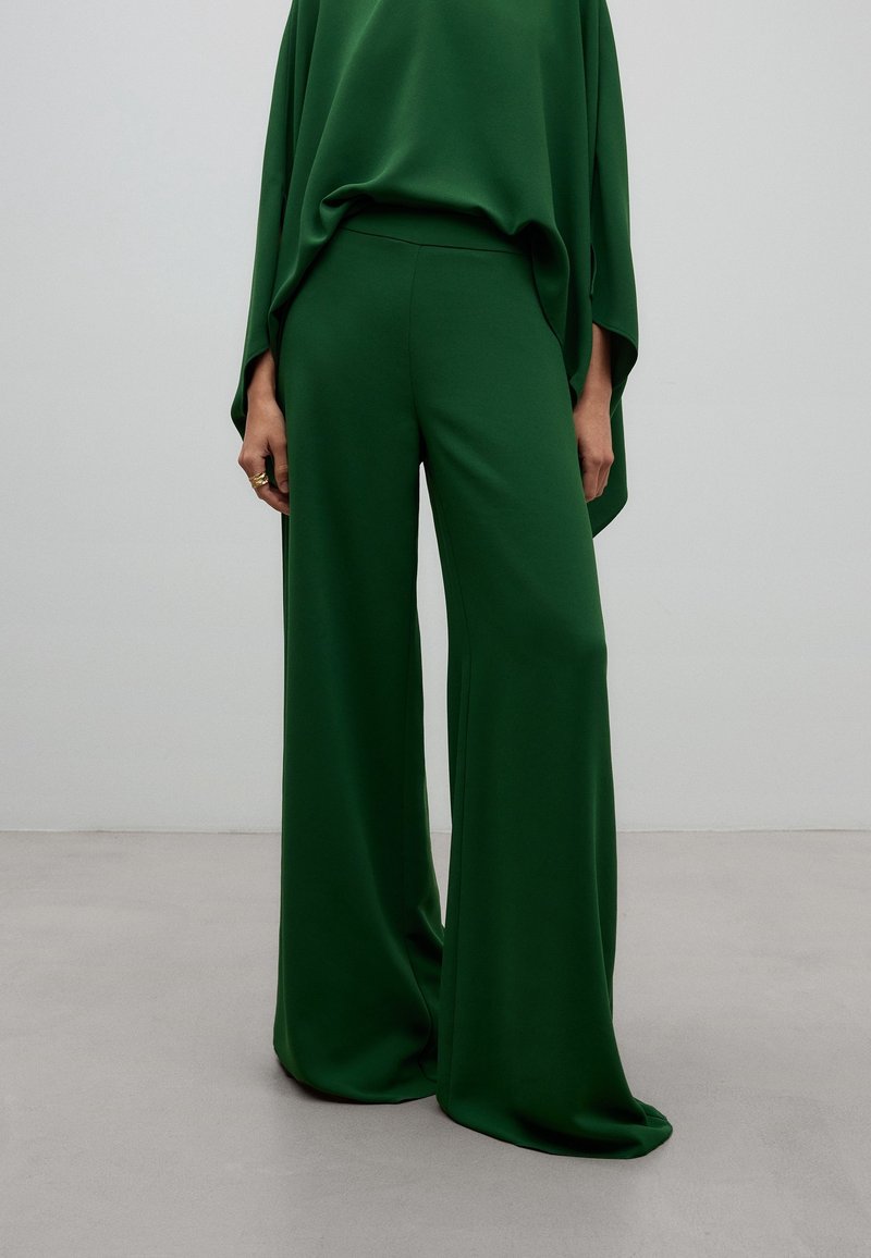 Femme portant un pantalon large taille haute vert foncé et un haut ample assorti, debout devant un fond clair uni.