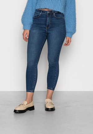 Persona que lleva vaqueros pitillo azules recortados, un suéter peludo azul claro y mocasines beige con suelas negras, de pie en un suelo gris.