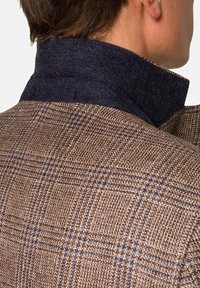 Homme portant une veste à carreaux marron avec un col relevé bleu foncé, vu de dos et de profil.