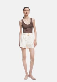 Bruine geribde tanktop gecombineerd met crèmekleurige shorts met een riem en gouden gesp. Model draagt strappy witte hakken. Eenvoudige achtergrond.