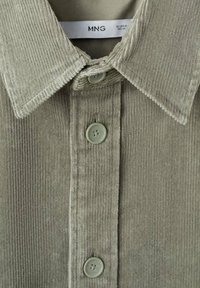 Camasa din corduroy verde, cu guler clasic, textură cu nervuri verticale și butoane de culoare deschisă, având un etichetă pe care scrie "MNG."
