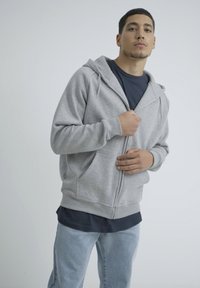 Gray zip-up hoodie with a front pocket, worn over a dark t-shirt, paired with light blue denim jeans. Smooth texture and casual design.