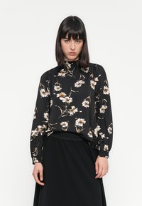 Kvinne med mørkt hår iført en svart bluse med høy hals med beige og hvite blomster og svart skjørt, står mot en hvit bakgrunn.