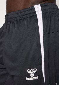 Mustad mustad lühikesed püksid, millel on külgedel valge vertikaalne triip ja logo alumisel säärel, on valmistatud kerge kangaga, millel on sile tekstuur.