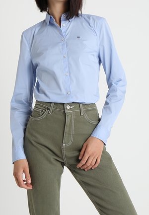 Femme portant une chemise bleu clair à boutons rentrée dans un pantalon vert olive taille haute, debout avec une main posée sur la cuisse.