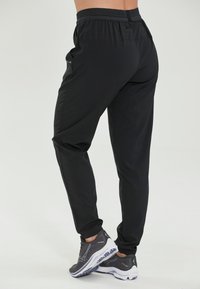 Sorte atletiske bukser med en tætsiddende design, der har en elastisk talje og sidelommer. Siddende sammen med grå sneakers med hvide såler.