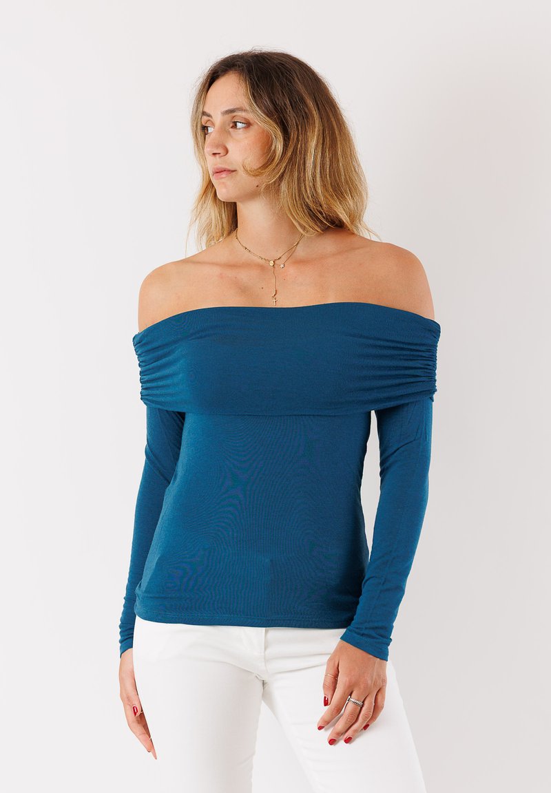 Top a maniche lunghe scivolato sulle spalle in un tessuto teal profondo, caratterizzato da una trama a coste e dettagli arricciati sul busto. Abbinato a pantaloni bianchi.