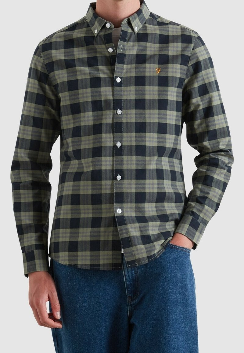 Homme portant une chemise à manches longues boutonnée avec un motif à carreaux vert et noir et un logo jaune brodé sur la poitrine, associée à un jean bleu.