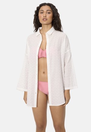 Mujer con una camisa blanca de ojales abotonada sobre un bikini rosa, de pie frente a un fondo gris claro liso.