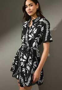 Robe à manches courtes noir et blanc avec un motif floral, ceinture à nouer et tissu texturé. Présente des accents brodés et une jupe évasée.