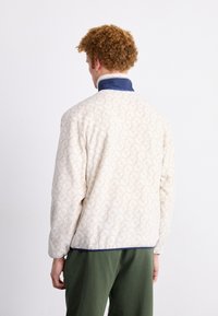 Chaqueta crema acolchada con un patrón sutil, cuello azul marino y tejido texturizado, diseñada para la comodidad y la versatilidad.