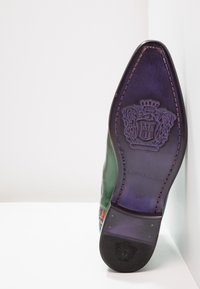 Suela de zapato de cuero verde con punta afilada y logotipo en relieve. Una tela colorida acentúa el costado, contrastando con la parte inferior morada.