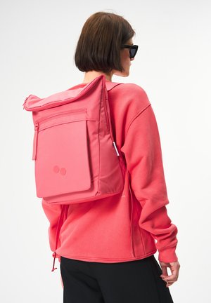 Rosa Stoff-Rucksack mit Roll-Top-Design, ausgestattet mit einer Front-Reißverschlusstasche und runden Akzenten. Wird auf dem Rücken über einem passenden Sweatshirt getragen.