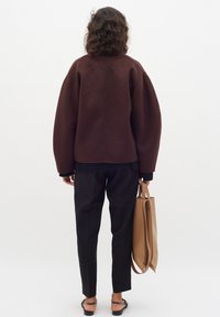 Rudas perteklinis megztinis su nuleistais petimi, derinamas su juodomis prigludusiomis kelnėmis. Bežinė rankinė laikoma rankoje, modelis nugarą atsukęs kamerai.