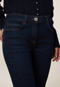 Jeans de denim azul oscuro con un diseño ajustado, que presentan costuras naranjas, un cierre de botón plateado y bolsillos frontales. Textura suave.