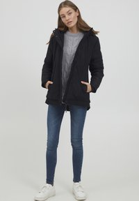 Svart polstret jakke med hette, glidelås foran og sidelommer, lagd over en grå kabelstrikket genser og blå skinny jeans, kombinert med hvite joggesko.