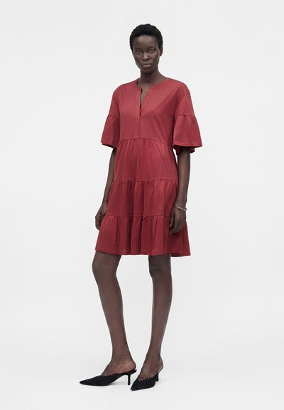 Γυναίκα με στρωτό, κόκκινο φόρεμα μέχρι τα γόνατα με κοντά μανίκια και άνοιγμα στο ντεκολτέ, φορόντας μαύρες μυτερές γόβες, στέκεται μπροστά σε απλό φόντο.