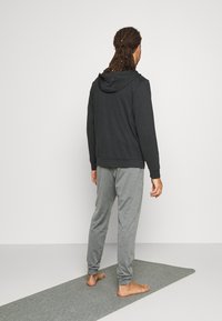 Sudadera gris con cremallera y capucha ajustable, combinada con joggers gris claro. Descalzo sobre una esterilla de yoga gris, de pie frente a un fondo sencillo.