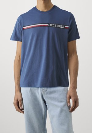 Mężczyzna w niebieskiej koszulce z krótkim rękawem, z poziomym paskiem w kolorach czerwonym, białym i czarnym oraz napisem "HILFIGER" na piersi.
