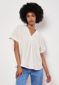 Blusa di colore crema a maniche corte con scollo a V e dettaglio plissettato sul davanti. Realizzata in un tessuto leggero e testurizzato. Vestibilità casual.