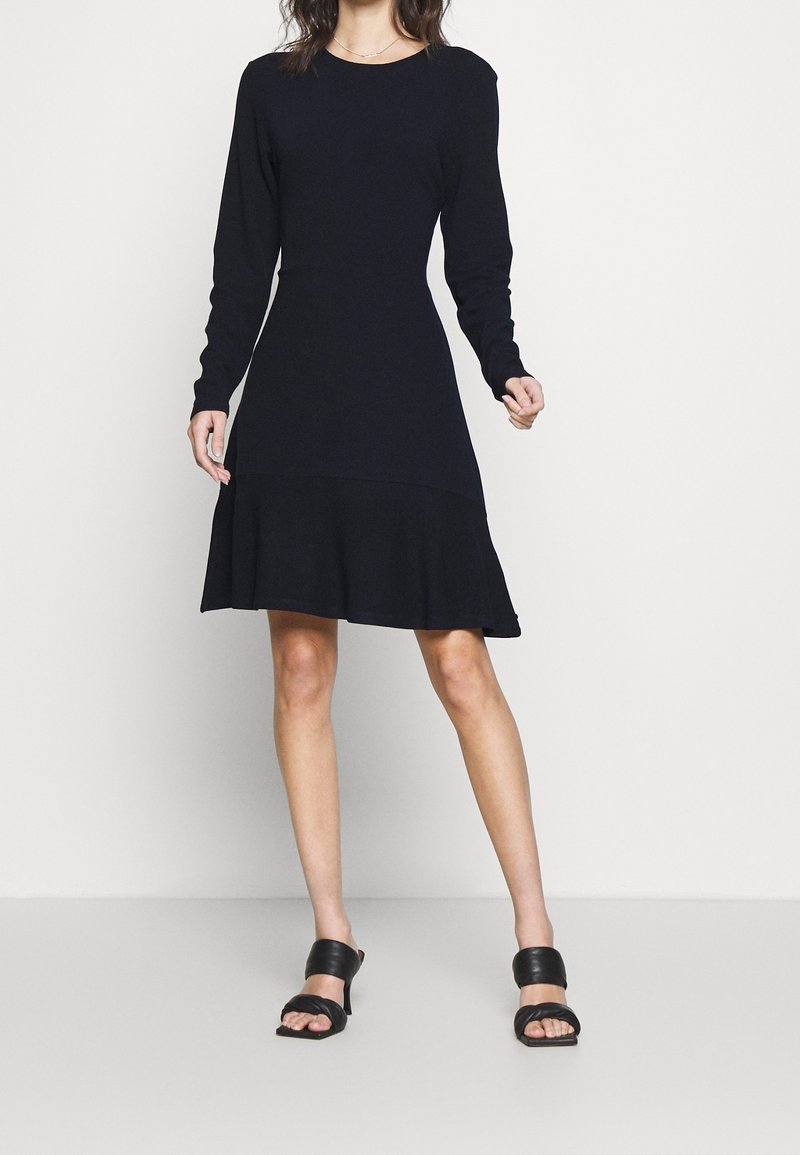 Femme portant une robe noire à manches longues, longueur genou, avec un ourlet évasé et des sandales noires à bouts ouverts, debout devant un fond blanc.