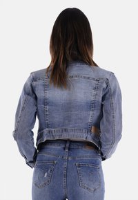 Giacca di jeans, lavaggio medio, design cropped con due tasche posteriori, dettagli in pelle marrone e una leggera scoloritura. La vista posteriore mostra i dettagli delle cuciture.