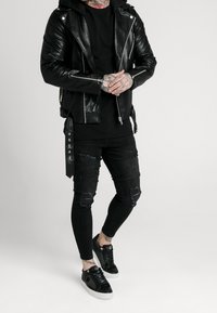 Czarna skórzana kurtka motocyklowa ze srebrnymi zamkami błyskawicznymi, czarna koszula z długim rękawem, podarte czarne obcisłe dżinsy oraz czarne trampki z białą podeszwą.