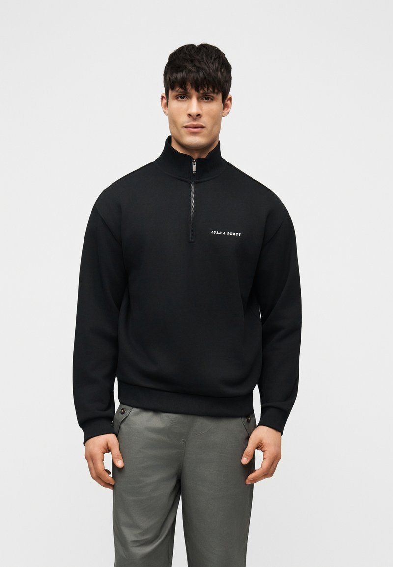 Άνδρας που κοιτάζει μπροστά, φοράει μαύρο φόρεμα με φερμουάρ στο ένα τέταρτο και λογότυπο «Lyle & Scott» και γκρι παντελόνια, σε ένα απλό ανοιχτό φόντο.