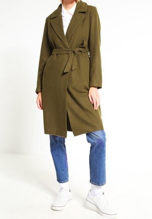 Mannequin portant un manteau ceinturé vert olive sur une chemise blanche, un jean bleu, des chaussettes blanches et des baskets blanches, debout devant un fond blanc uni.