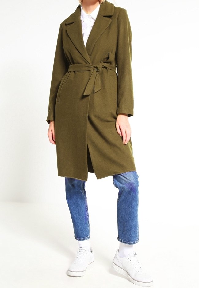 Mannequin portant un manteau ceinturé vert olive sur une chemise blanche, un jean bleu, des chaussettes blanches et des baskets blanches, debout devant un fond blanc uni.