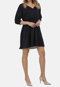 Schwarzes, tailliertes Kleid mit Dreiviertelärmeln, V-Ausschnitt und schwingendem Rock. Kombiniert mit nude-farbenen High Heels.