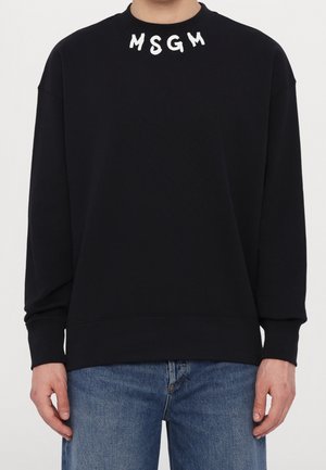 Czarna bluza z długim rękawem z białym napisem „MSGM” tuż przy dekolcie, noszona z niebieskimi dżinsami, widoczna od klatki piersiowej do bioder.