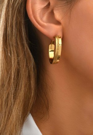 Boucle d'oreille créole en or avec des motifs texturés et lisses sur la moitié, portée à l'oreille gauche d'une personne aux cheveux châtain clair visibles.