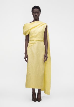 Femme portant une robe jaune texturée une épaule avec une longue manche drapée, associée à des talons noirs, debout sur un fond blanc.