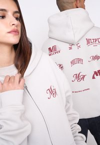 Svetlošedá mikina na zips s červenými grafickými logami na chrbte a prednej strane. Mäkký materiál s voľným strihom a šnúrkou na kapucni.
