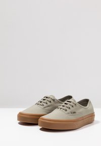 Baskets en toile beige à tige basse avec des semelles et des lacets en caoutchouc marron, placées côte à côte sur une surface blanche.