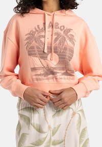 Sudadera color coral con un gráfico frontal que presenta palmeras y un sol naciente, capucha con cordón y corte relajado, combinada con pantalones cortos ligeros con estampado.