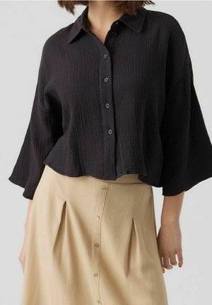 Kobieta w luźnej czarnej koszuli na guziki z rękawami do połowy przedramienia oraz beżowej spódnicy na guziki z plisami.