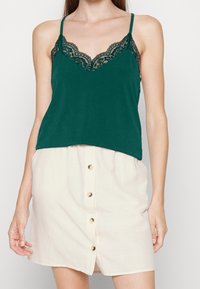 Top verde con pizzo e spalline regolabili, abbinato a una gonna chiara beige con bottoni realizzata in un morbido tessuto testurizzato.
