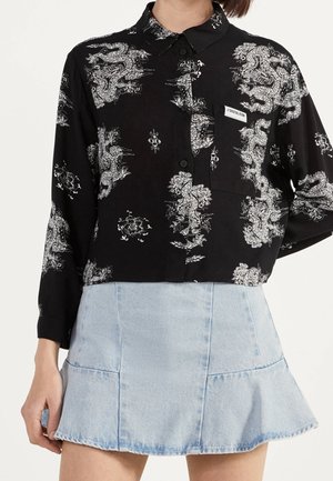 Zwarte blouse met knopen en witte draak- en bloemenpatronen, gecombineerd met een lichtblauwe denim minirok met een licht uitlopende zoom.